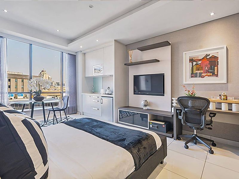 Modern bedroom featuring a compact built-in kitchenette, floor-to-ceiling windows with city views, recessed lighting, and sleek tiled flooring.
