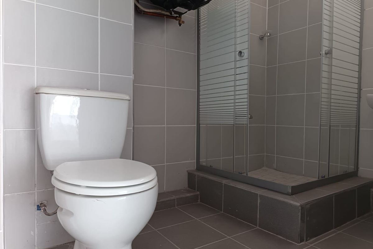 A functional bathroom featuring a tiled shower enclosure, toilet, and wall-mounted electric water heater with neutral grey tiling throughout.