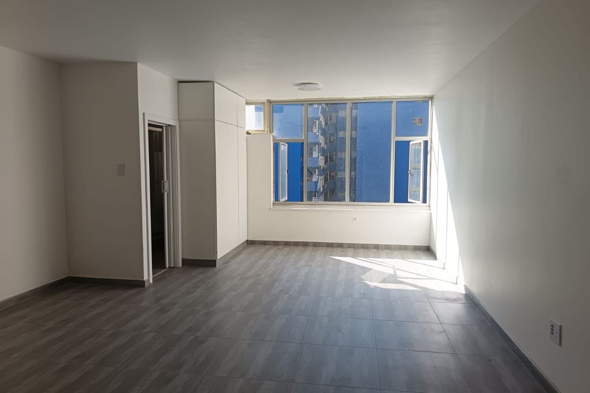 A bright, empty living area featuring modern grey tiled flooring, built-in storage cupboards, and large windows providing natural light.