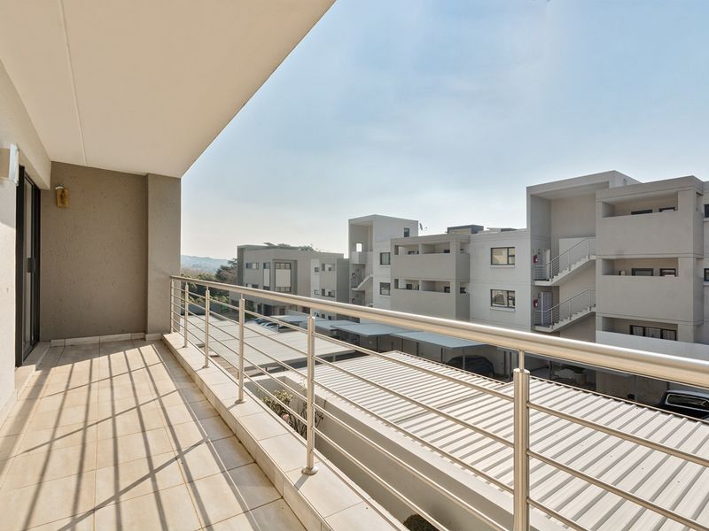 A spacious, covered balcony featuring tiled flooring and a modern stainless steel balustrade, offering an elevated view of the apartment complex.