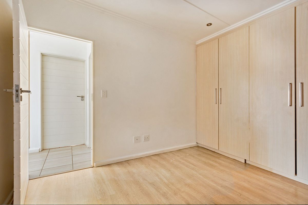A bright, empty bedroom featuring light wood-look laminate flooring and ample built-in cupboard storage, leading into a tiled hallway.