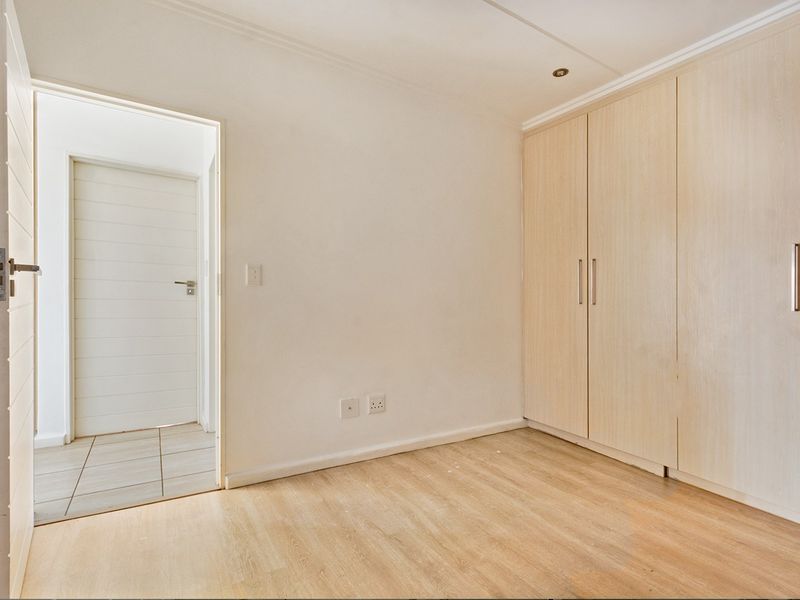 A bright, empty bedroom featuring light wood-look laminate flooring and ample built-in cupboard storage, leading into a tiled hallway.