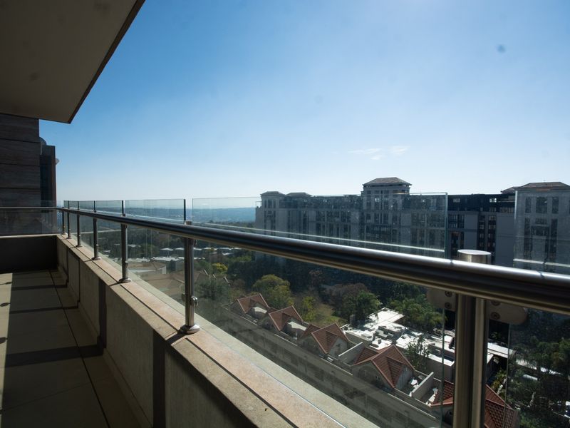 A modern balcony featuring a sleek glass balustrade with a stainless steel handrail, offering expansive elevated views of the surrounding urban landscape.