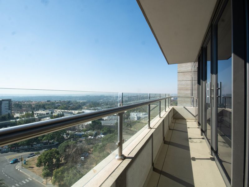 A modern apartment balcony featuring a glass balustrade with stainless steel handrails, tiled flooring, and expansive views of the surrounding area.
