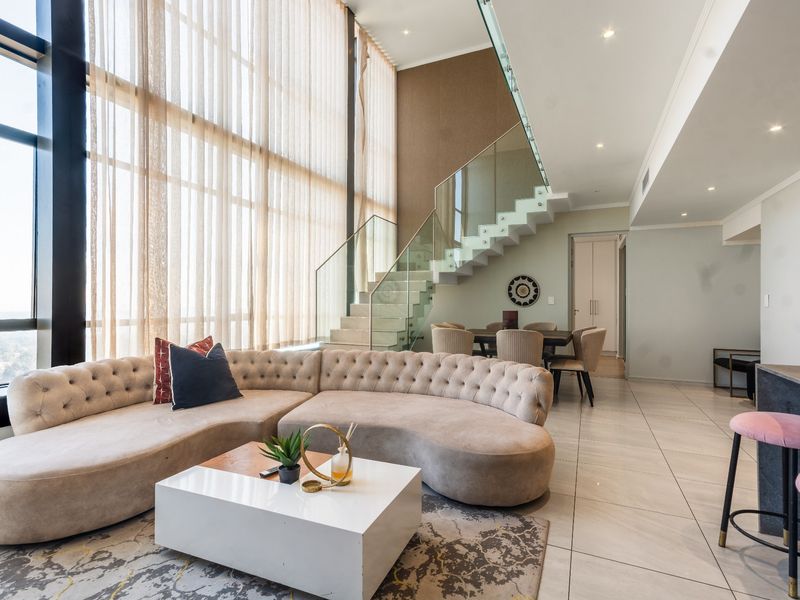 Spacious open-plan living area featuring double-volume windows, modern tiled flooring, and a striking glass balustrade staircase leading to the upper level.