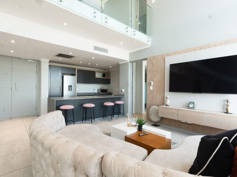 Modern open-plan living area featuring a sleek kitchen with island, double-volume ceilings, glass mezzanine balustrade, and integrated media wall.