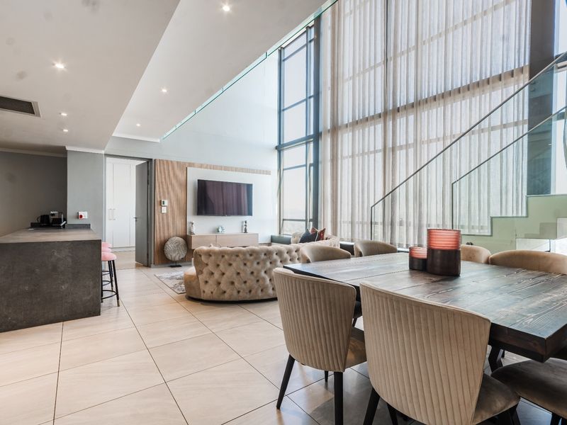 Spacious open-plan living and dining area featuring double-height windows, a modern kitchen island, and a sleek glass staircase in a contemporary apartment setting.