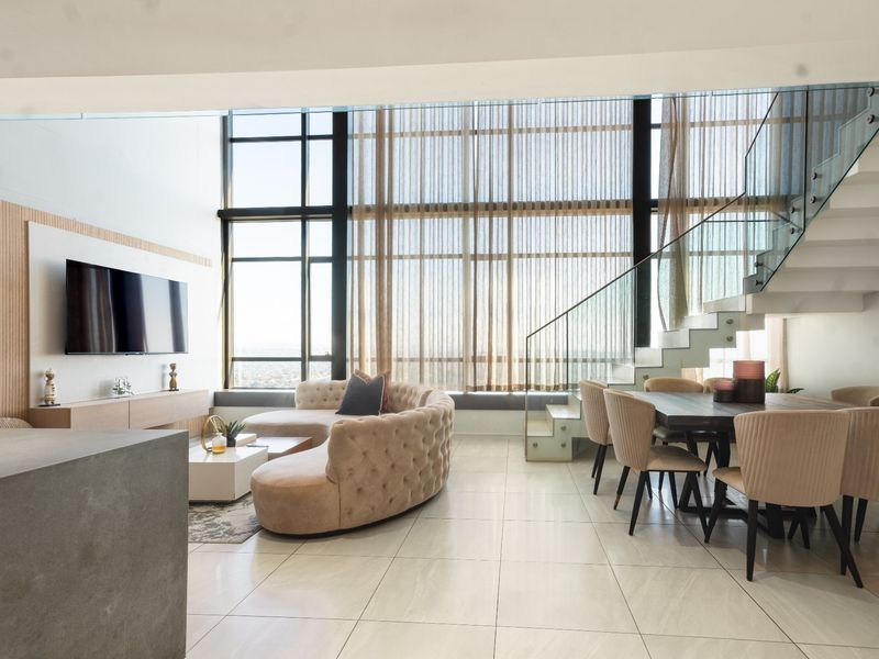 Spacious open-plan living and dining area featuring double-volume windows, modern tiled flooring, and a striking glass balustrade staircase.