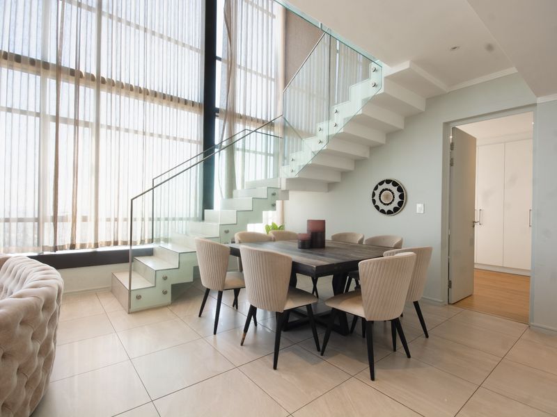 Modern open-plan living and dining area featuring a striking glass staircase, double-volume floor-to-ceiling windows, and sleek tiled flooring.