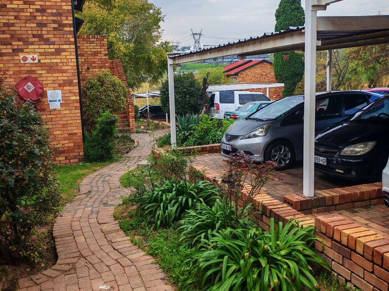 Exterior view of a residential apartment complex featuring a brick-paved walkway, landscaped garden beds, and a covered carport area.