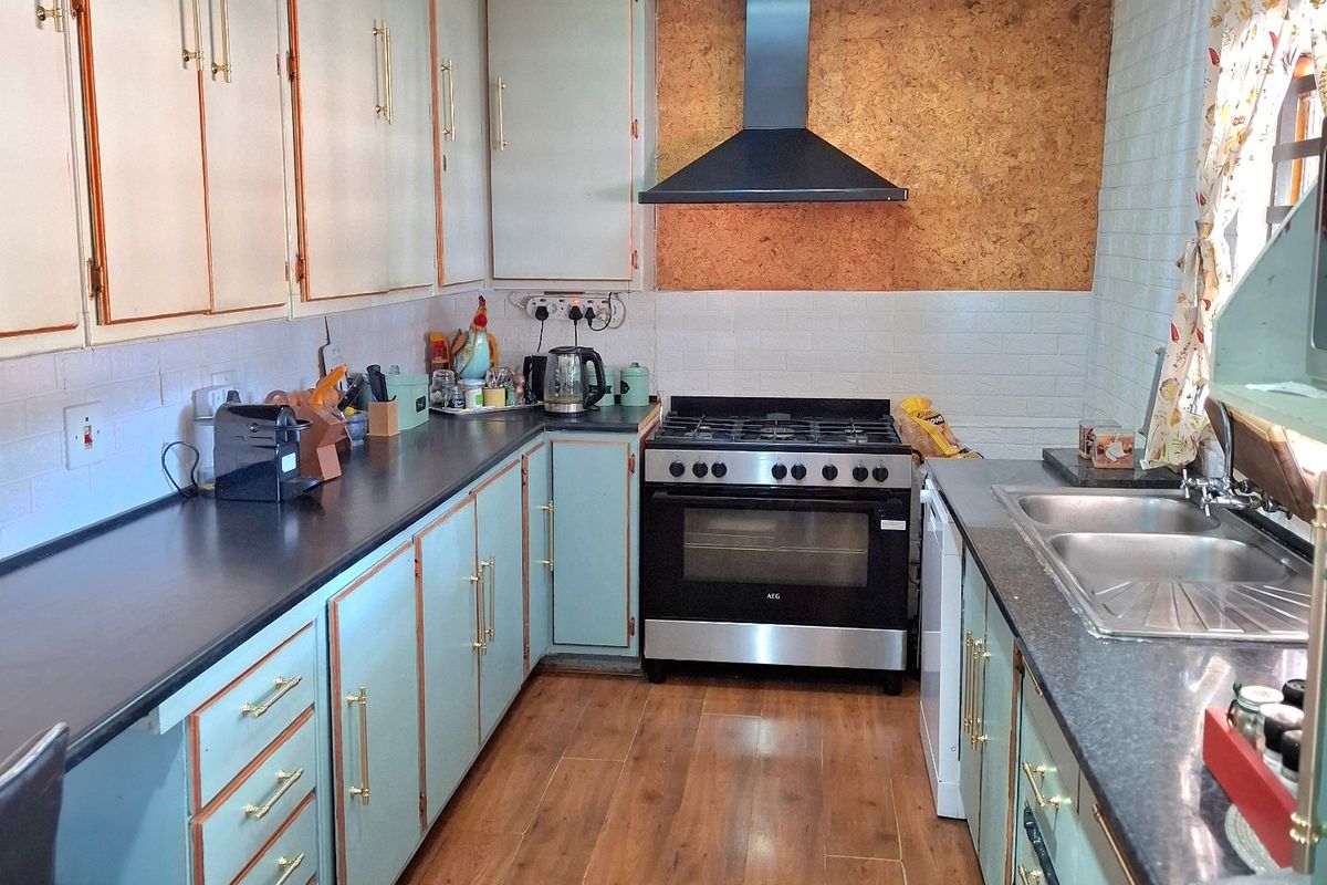 A functional galley-style kitchen featuring light-colored cabinetry, dark countertops, a freestanding oven with extractor fan, and wood-look laminate flooring.
