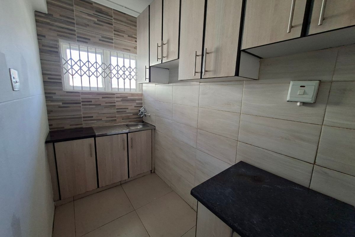 A compact, functional kitchen featuring light wood-tone cabinetry, dark countertops, tiled splashbacks, and a window with security bars.