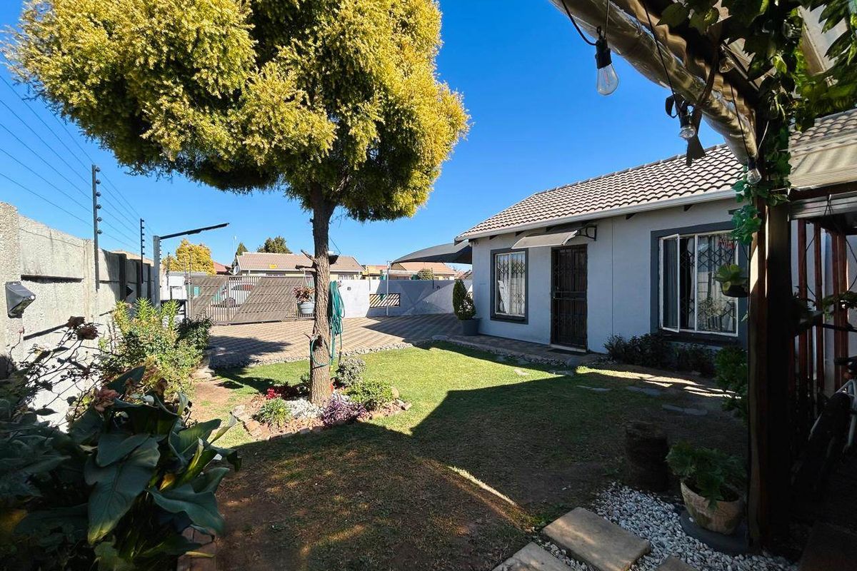 A well-maintained rear exterior view featuring a paved driveway, a small lawn area, and secure perimeter fencing with security bars on windows.