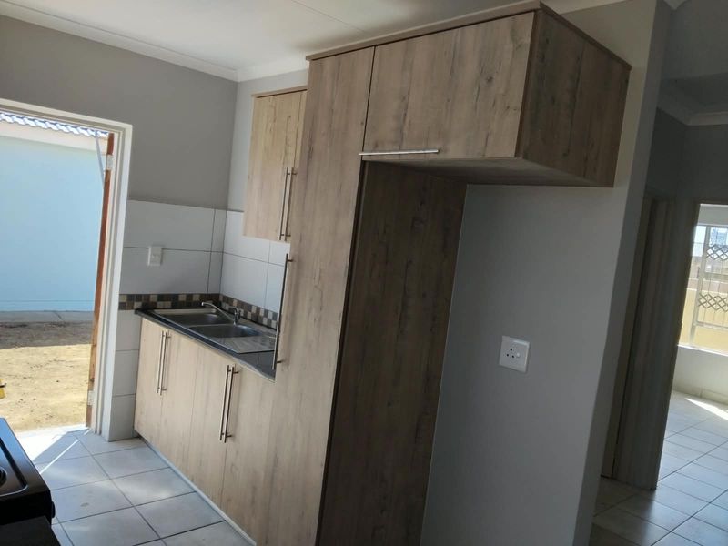 A clean, modern kitchen featuring wood-look cabinetry, laminate countertops, a stainless steel double sink, and neutral tiled flooring with direct outdoor access.