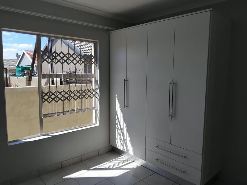 A bright, empty bedroom featuring modern white built-in wardrobes, tiled flooring, and a window fitted with a security gate.