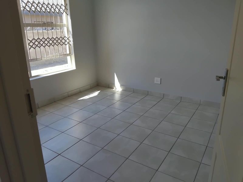 A clean, empty bedroom featuring neutral tiled flooring, a window with a security gate, and a simple, well-maintained interior finish.