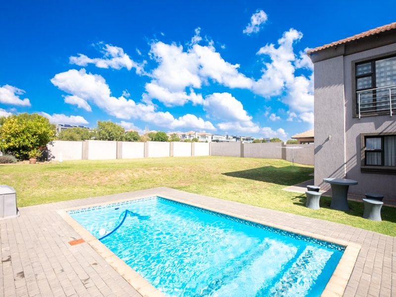 A well-maintained backyard featuring a swimming pool with paved surround, expansive lawn, and perimeter walling, set against a clear blue sky.