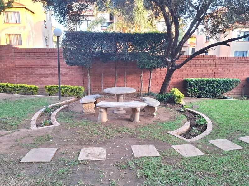 A communal garden area featuring a stone table and bench set, surrounded by established trees, manicured hedges, and a brick boundary wall.