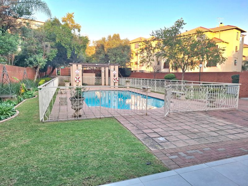 Communal pool area featuring a paved deck, metal safety fencing, and a pergola, set within a landscaped garden with brick boundary walls.