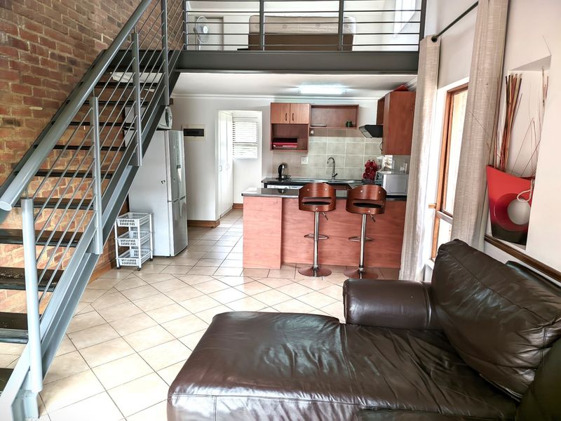 Open-plan living area featuring a mezzanine level, exposed brick wall, metal staircase, and a functional kitchen with granite countertops and wood-finish cabinetry.