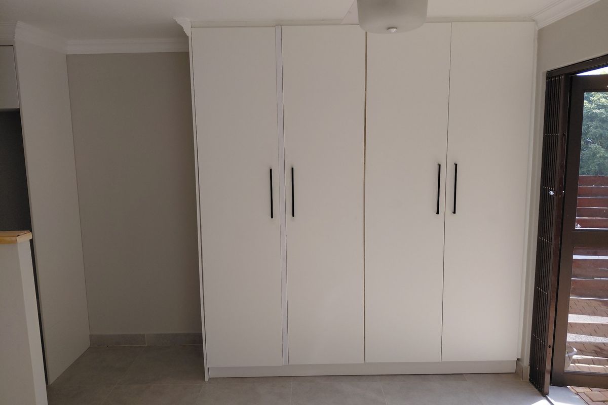 A clean, empty bedroom featuring modern white built-in cupboards, neutral tiled flooring, a ceiling fan, and direct access to an outdoor area via a security gate.