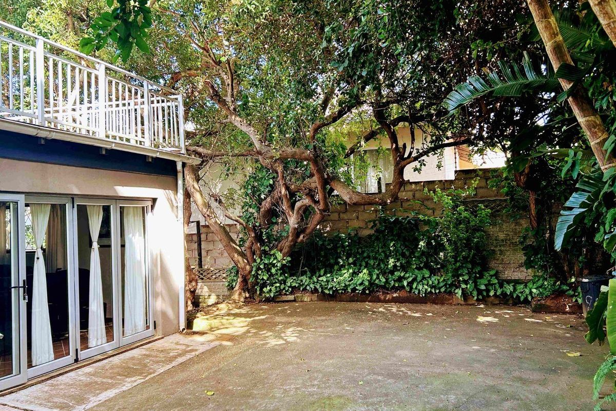 A rear exterior view featuring a concrete patio area, mature trees, a retaining wall, and a balcony with metal railings attached to the house.