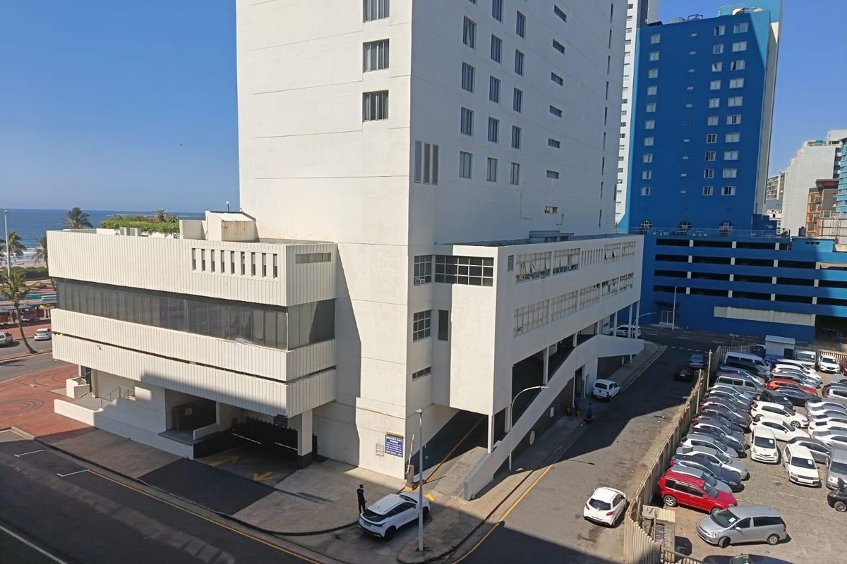An elevated aerial view of a multi-story apartment building featuring a parking ramp and adjacent parking area in an urban coastal setting.