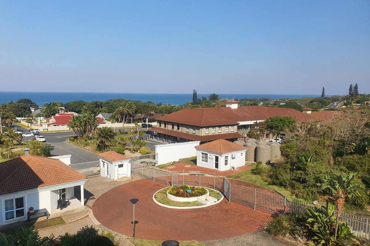 Aerial view of the apartment complex grounds, featuring a paved driveway, security entrance, and landscaped gardens with a distant ocean view.