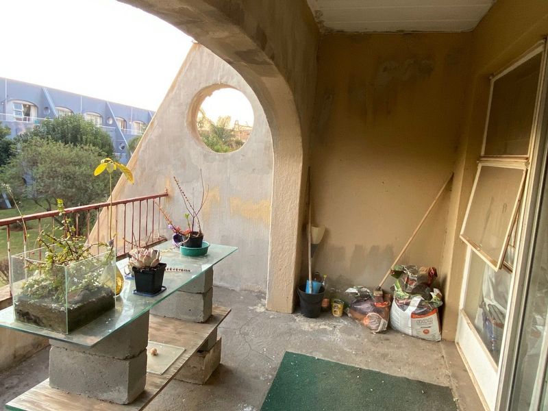 A covered balcony featuring an arched wall with a circular opening, metal railing, and sliding glass door access, requiring cosmetic maintenance and surface repairs.