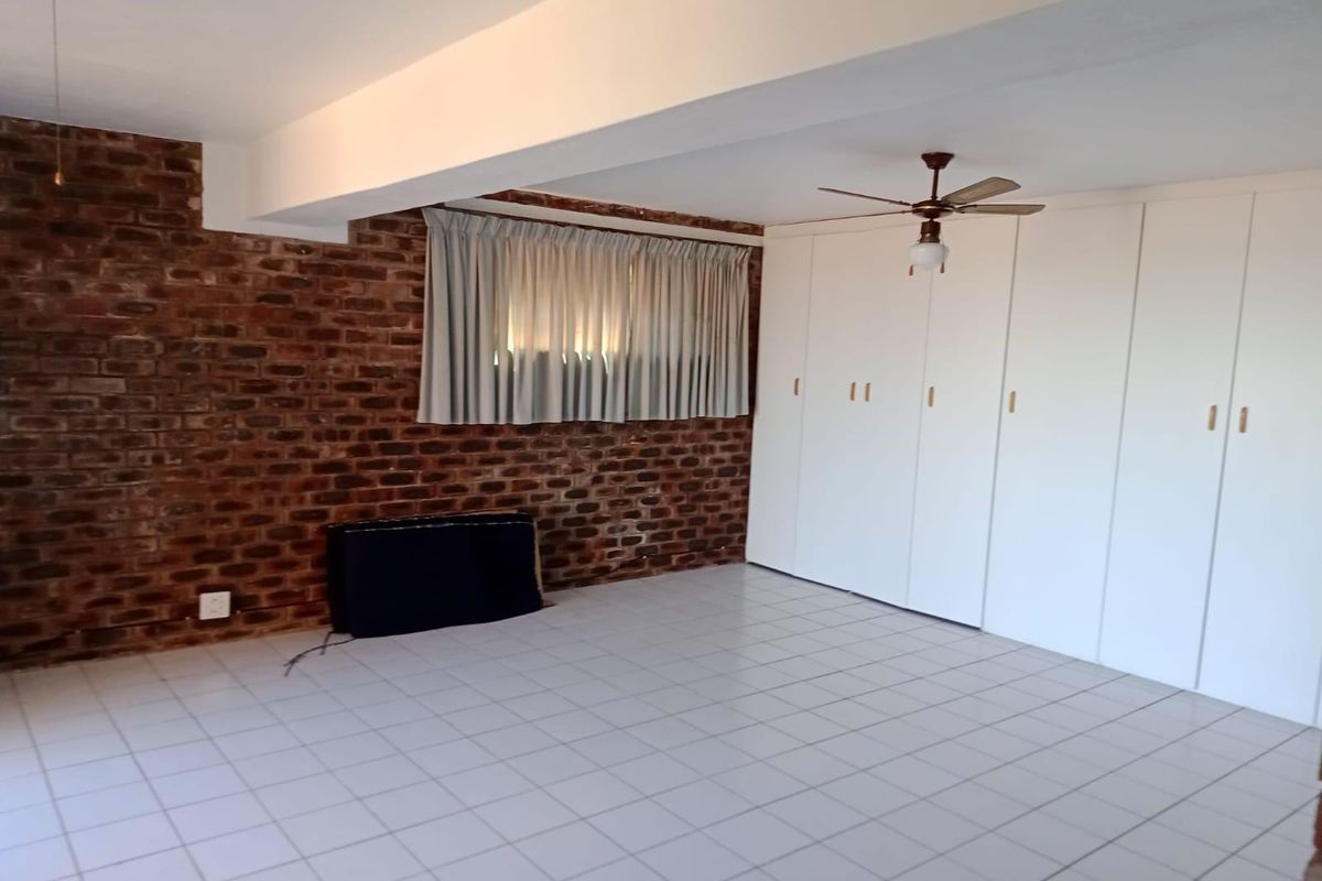 Spacious bedroom featuring an exposed brick feature wall, extensive built-in cupboards, a ceiling fan, and neutral tiled flooring.