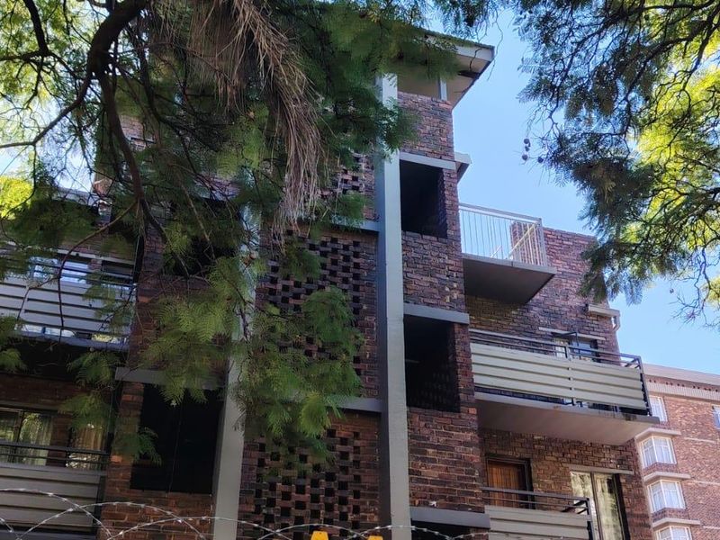 Exterior view of a multi-story apartment building featuring face brick construction, private balconies with metal railings, and established surrounding trees.