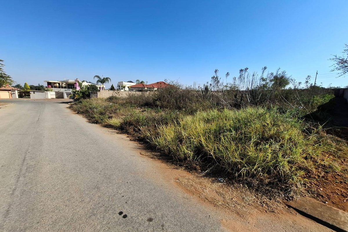 A street-level view of a vacant residential plot featuring natural vegetation and adjacent paved road access in a developed neighborhood.