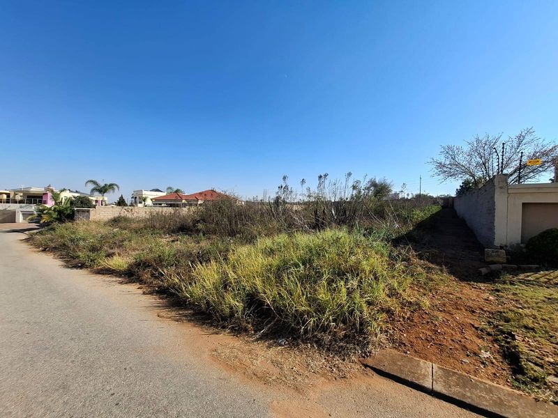 A vacant residential plot featuring boundary walls with electric fencing, accessible via a paved road in a developed neighborhood.