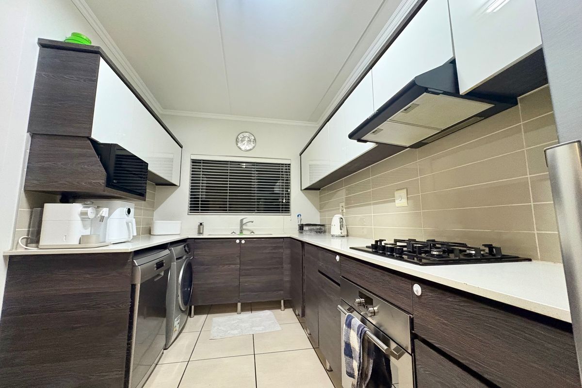 Modern galley-style kitchen featuring dark wood-grain cabinetry, white high-gloss upper cabinets, gas hob, built-in oven, and integrated laundry appliance space.