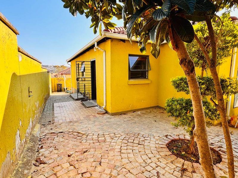 A rear exterior view of the property featuring a paved courtyard, security gate, and yellow painted exterior walls with some visible wear.