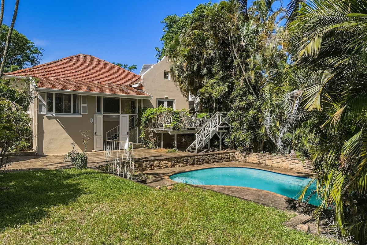 Rear view of the property featuring a swimming pool, stone retaining wall, and an elevated wooden deck with stairs leading to the garden.