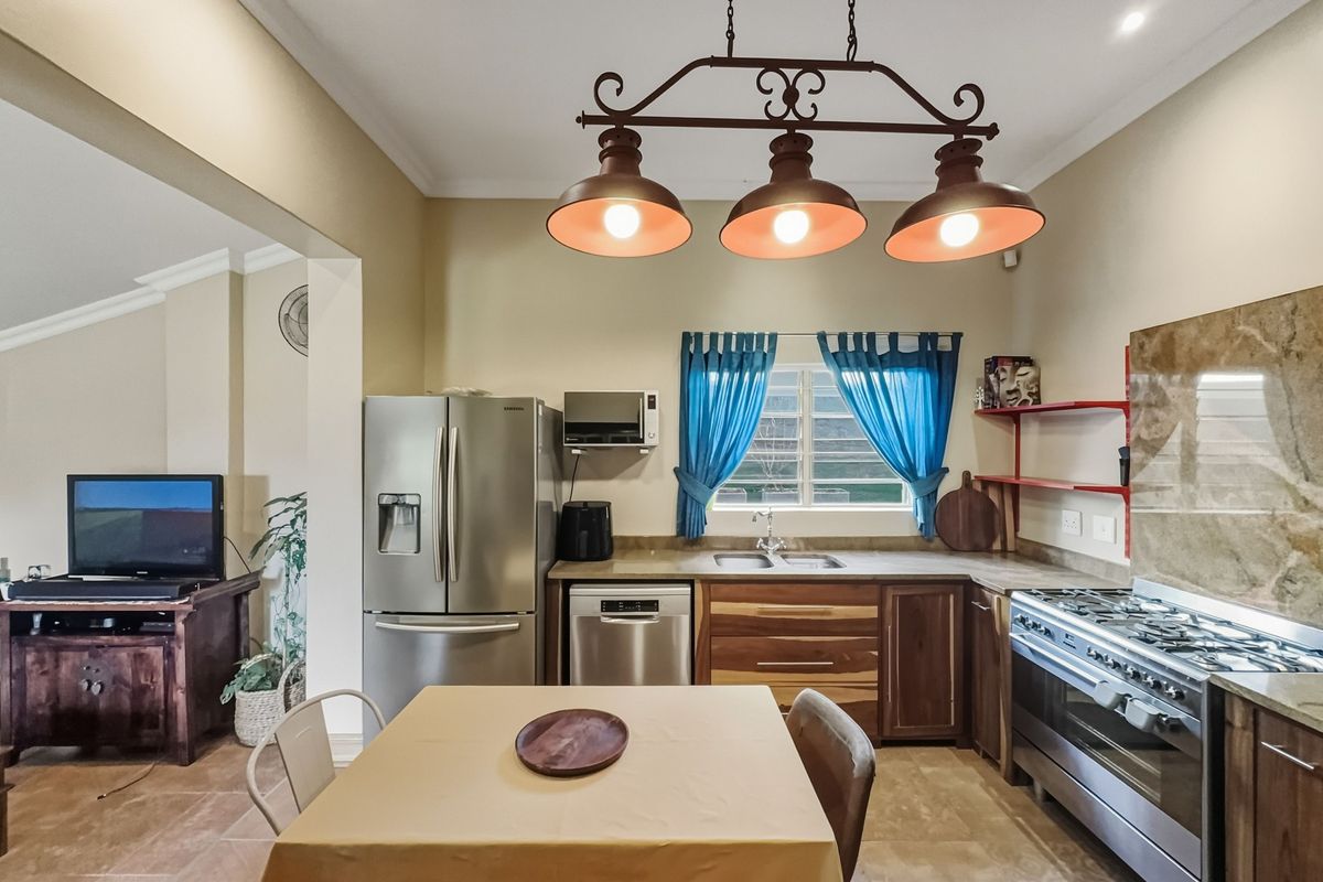 A functional kitchen featuring wooden cabinetry, a gas stove with oven, stone splashback, and a double sink, illuminated by a triple pendant light fixture.
