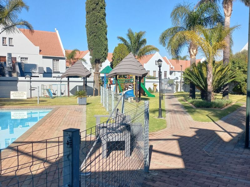 Communal pool area featuring a paved walkway, mesh safety fencing, and a small playground set within a landscaped garden environment.