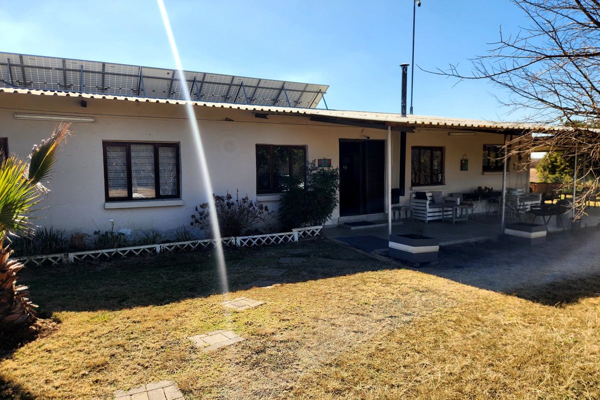 Rear view of the farmhouse featuring a covered patio, solar panel installation on the roof, and a surrounding garden area.