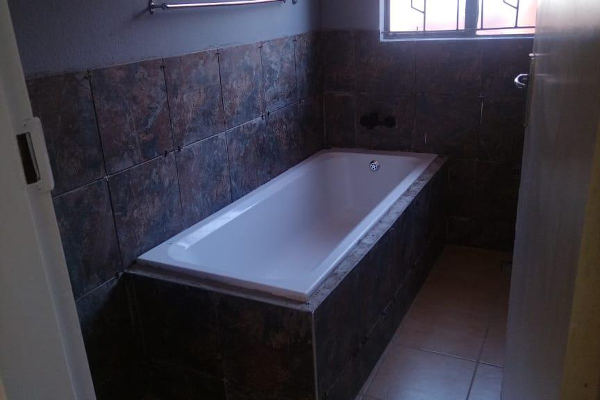 A functional bathroom featuring a built-in bathtub with dark tiled surround, tiled flooring, and a window with security bars.