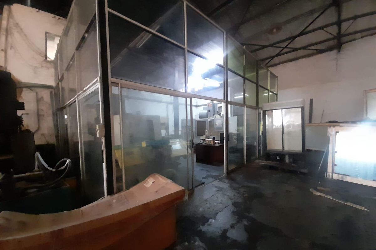 Industrial warehouse space featuring a glass-partitioned office enclosure, exposed steel roof trusses, and concrete flooring, suitable for manufacturing or workshop operations.
