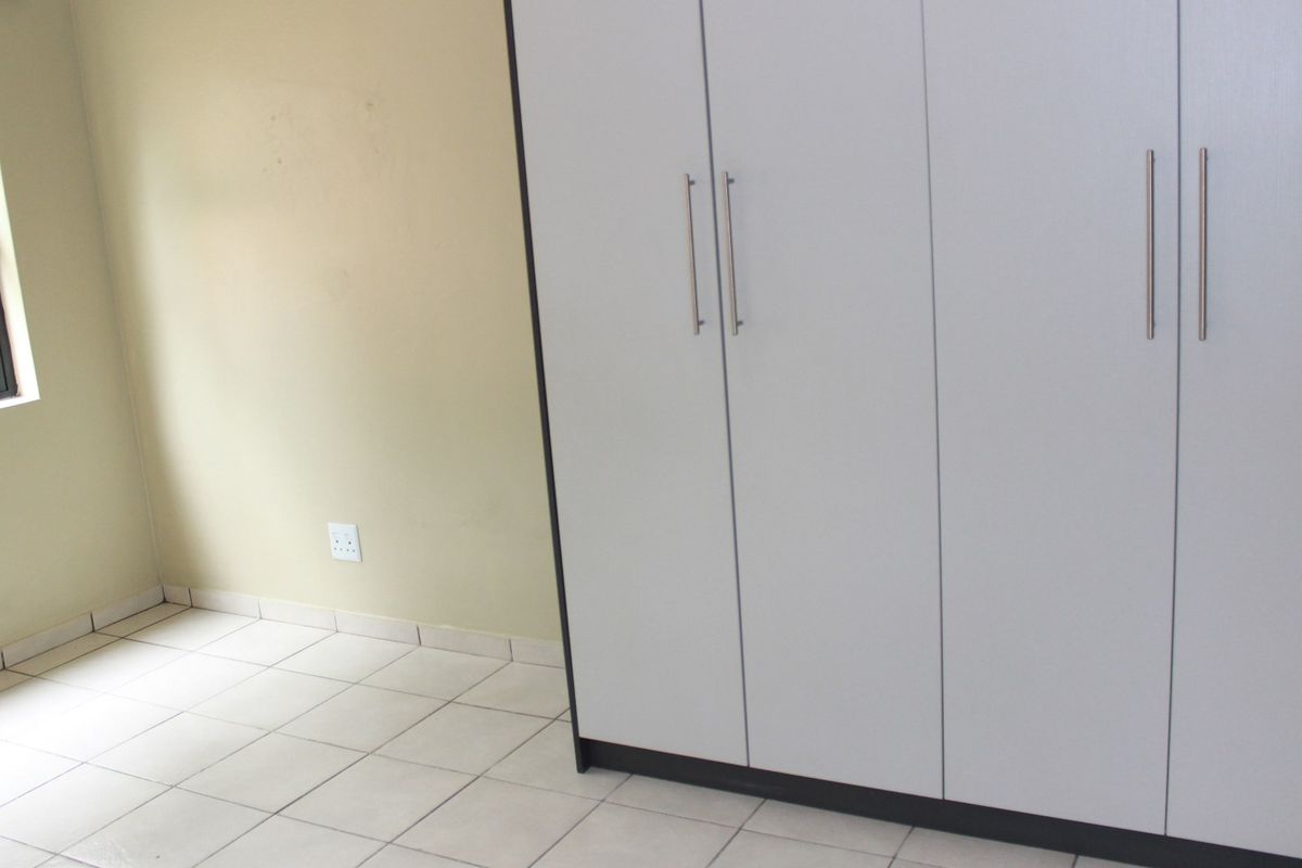 A bright, empty bedroom featuring neutral tiled flooring, a large built-in wardrobe with modern handles, and a window providing natural light.