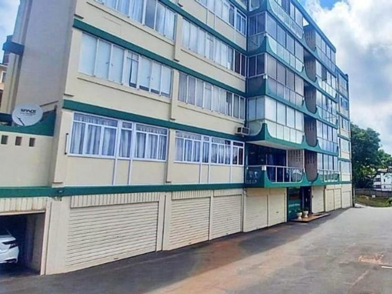 Exterior view of a multi-story apartment building featuring enclosed balconies, roller shutter garage doors, and a paved driveway area.