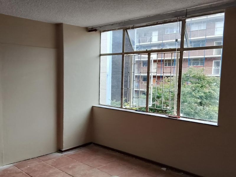 An empty bedroom featuring large steel-framed windows, tiled flooring, and a built-in cupboard, requiring cosmetic updates to improve its overall condition.