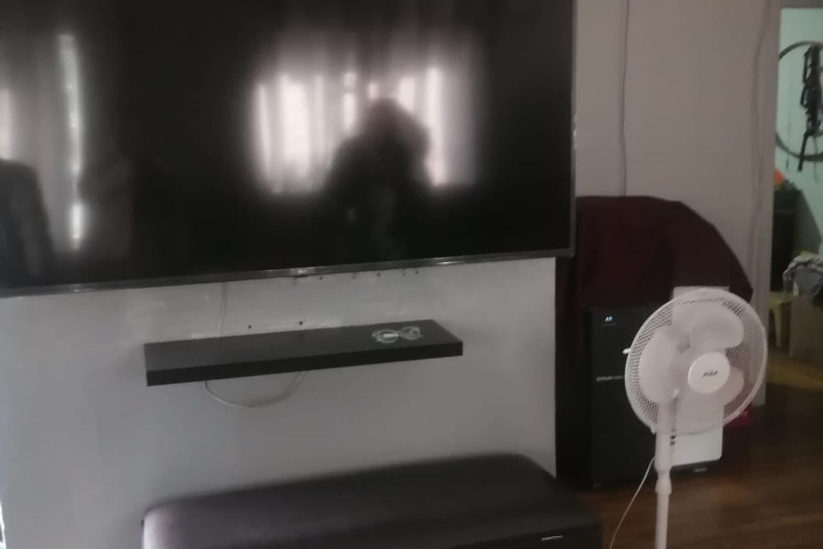 A living room area featuring hardwood flooring and a wall-mounted shelf, currently showing signs of wear and exposed wiring.