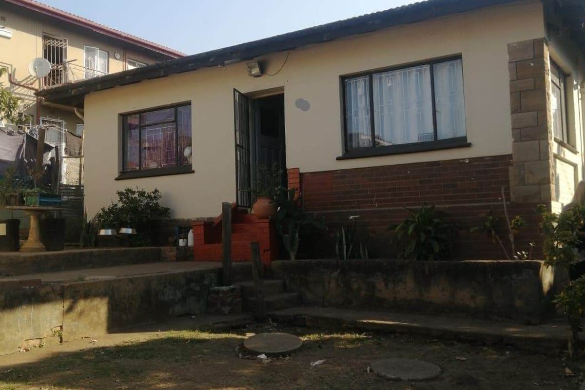 Rear view of a single-story house featuring a brick and plaster facade, concrete retaining wall, and steps leading to the entrance.
