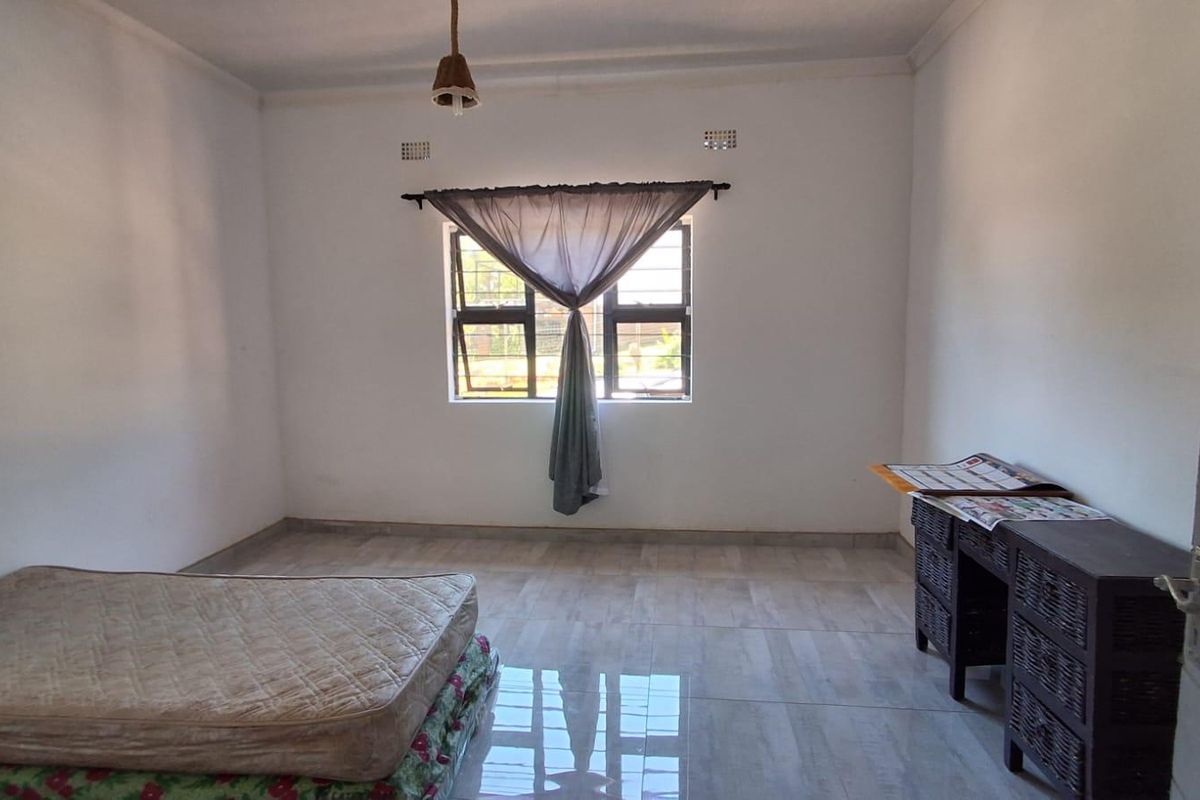 A simple, neutrally painted bedroom featuring tiled flooring, a window with security bars, and a central ceiling light fixture.