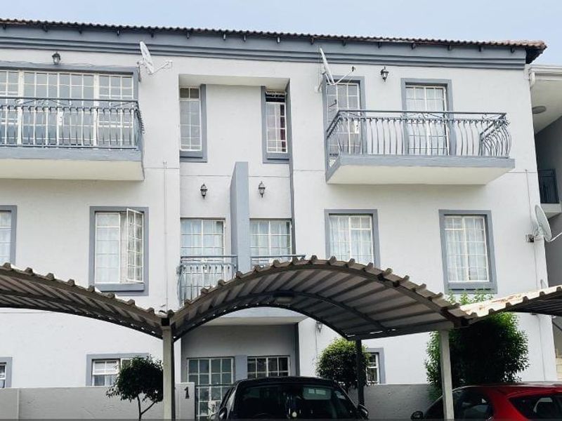 Front elevation of a multi-story apartment complex featuring balconies with wrought iron railings and covered parking bays.
