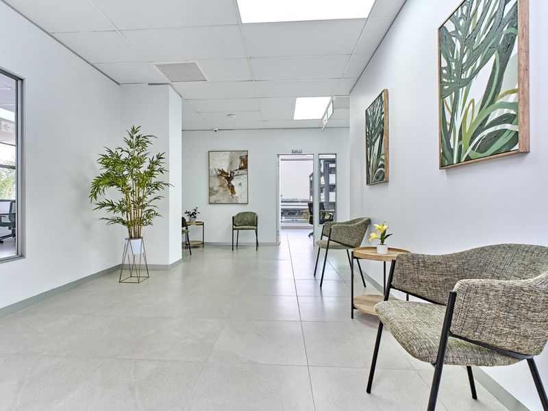 A bright, modern commercial hallway featuring neutral tiled flooring, a suspended ceiling with recessed lighting, and a glass partition wall.