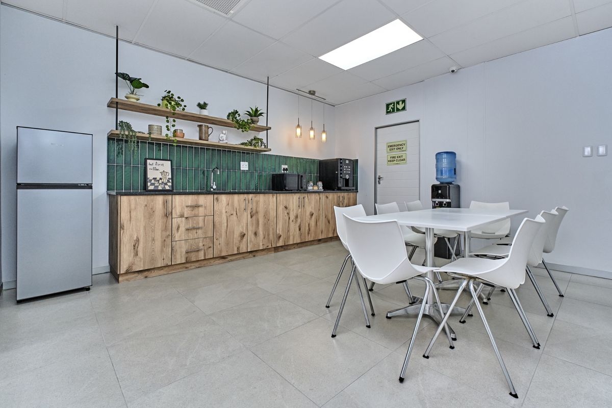A modern commercial kitchenette featuring wood-grain cabinetry, a green tiled backsplash, floating shelving, and a dedicated dining area with tiled flooring.